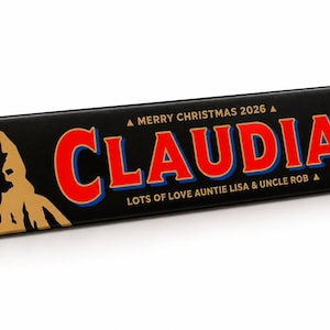 Pu&ograve; includere: Una scatola triangolare di cioccolato Toblerone con involucro nero. La parte anteriore presenta il nome CLAUDIA in grandi lettere rosse con contorni blu, il testo MERRY CHRISTMAS 2026, LOTS OF LOVE AUNTIE LISA & UNCLE ROB e un'etichetta 25 DEC 2026.
