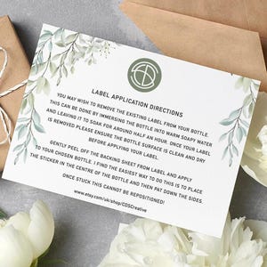 Puede incluir: Una tarjeta blanca con instrucciones de aplicaci&oacute;n de etiquetas, adornada con vegetaci&oacute;n de acuarela, descansa sobre una superficie gris. Tambi&eacute;n hay una caja de regalo marr&oacute;n envuelta con hilo y un sobre marr&oacute;n. El texto de la tarjeta incluye instrucciones.