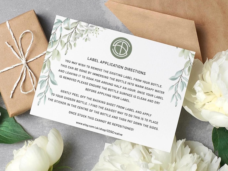 Puede incluir: Una tarjeta blanca con instrucciones de aplicaci&oacute;n de etiquetas, adornada con vegetaci&oacute;n de acuarela, descansa sobre una superficie gris. Tambi&eacute;n hay una caja de regalo marr&oacute;n envuelta con hilo y un sobre marr&oacute;n. El texto de la tarjeta incluye instrucciones.