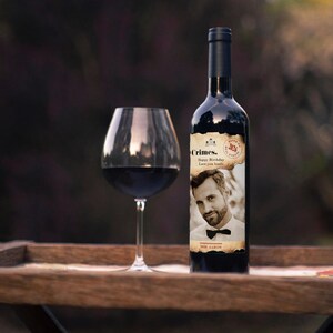 Puede incluir: Una botella de vino con una etiqueta personalizada con la foto de un hombre y el texto "Crimes. Happy Birthday Love you loads". Una copa de vino tinto est&aacute; junto a la botella en una bandeja de madera.