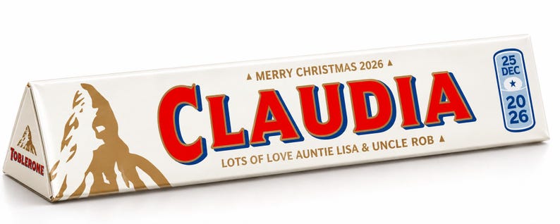 Pu&ograve; includere: Una confezione triangolare bianca di cioccolato Toblerone con il nome CLAUDIA in grandi lettere rosse. Il testo recita MERRY CHRISTMAS 2026, LOTS OF LOVE AUNTIE LISA & UNCLE ROB, e include un'icona di calendario con 25 DEC 2026.