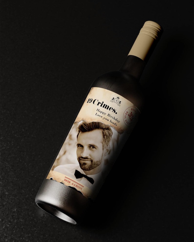 Puede incluir: Una botella de vino oscuro con una etiqueta crema que muestra la foto de un hombre y el texto "19 Crimes. Happy Birthday Love you loads." La botella tiene una base plateada y un cuello beige. Tambi&eacute;n aparece el nombre "Aaron".