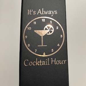 May include: Black leather-bound book with gold lettering that reads "It's Always Cocktail Hour". The cover features a gold clock face with a martini glass and a lemon slice.