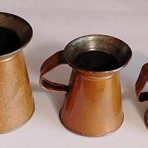 May include: A set of three graduated copper pitchers with dark metallic rims and handles. Each pitcher features a wide, flared opening that tapers down to a circular base. The exterior surfaces display a warm, polished copper finish with subtle variations in tone and texture across the three distinct sizes.