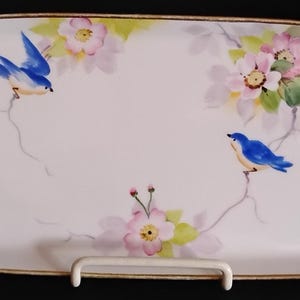 May include: An oblong white porcelain serving tray with gold-trimmed edges and integrated handles. The surface features a hand-painted design of two blue birds perched on thin branches surrounded by clusters of light pink flowers and green leaves.