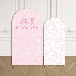 May include: Two arched backdrop covers featuring a white seashell pattern. The larger cover is light pink with the text She'll be here soon in pink script. The smaller cover is white with a light pink seashell pattern, both with rounded tops.