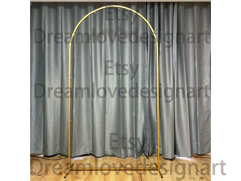 May include: A gold metal arch, suitable for weddings or events, is positioned in front of a grey curtain backdrop. The arch has a simple, elegant design, ideal for creating a focal point. The Etsy shop name Dreamlovedesignart is visible on the image.