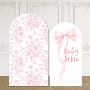 May include: Two arched backdrop panels. The taller left panel features a repeating light pink floral pattern on a white background. The shorter right panel is white and displays a large light pink ribbon bow above the text Baby Sofia.
