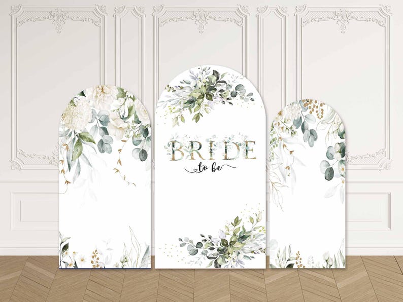 May include: Three white arched panels with floral and greenery designs. The centre panel reads "BRIDE to be" in gold lettering. The panels are decorated with white flowers, green leaves, and gold accents, ideal for a wedding or bridal shower.