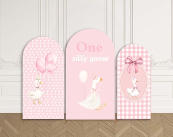 May include: Three pink arch backdrops with goose illustrations. The left backdrop has a polka dot pattern and a goose with balloons. The center backdrop says "One silly goose". The right backdrop has a gingham pattern and a goose with a bow.