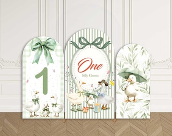 May include: Custom stretchy arch backdrop featuring three panels with a whimsical goose theme. The panels have a green and white color scheme, with the text "One Silly Goose" on the center panel. The backdrop is designed for a birthday party.