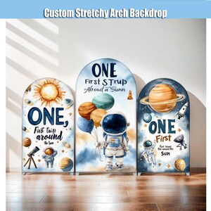 Puede incluir: Tres fondos de arco elásticos personalizados con temática espacial. Cada arco presenta la palabra "ONE" e ilustraciones relacionadas con el espacio, incluyendo planetas, cohetes y astronautas. El texto "Custom Stretchy Arch Backdrop" está en la parte superior.