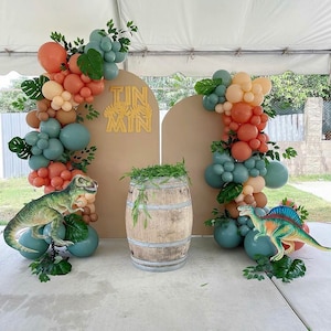 May include: A brown and tan backdrop with a gold 'Tin Man' sign.  The backdrop is decorated with a balloon arch of peach, beige, and teal balloons.  Two dinosaur figurines are on either side of a wooden barrel.