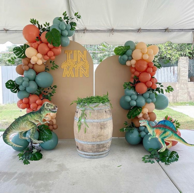 May include: A brown and tan backdrop with a gold 'Tin Man' sign.  The backdrop is decorated with a balloon arch of peach, beige, and teal balloons.  Two dinosaur figurines are on either side of a wooden barrel.