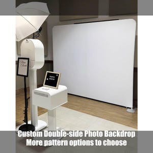 May include: A white photo booth setup with a large white backdrop. The booth has a sign that says "Photo Booth" and the text "Custom Double-side Photo Backdrop More pattern options to choose". A light and a small sign are also present.