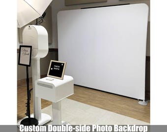 May include: A white photo booth setup with a large white backdrop. The booth has a sign that says "Photo Booth" and the text "Custom Double-side Photo Backdrop More pattern options to choose". A light and a small sign are also present.