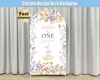 May include: Custom design arch backdrop with floral and watercolor elements. The backdrop features the text "Sophia is ONE" and a cartoon goose. The backdrop is framed by a floral border and has a fast turnaround.