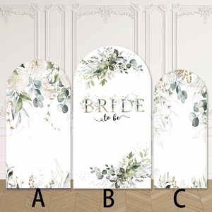 May include: Three white arched panels with floral designs and greenery. The central panel reads "BRIDE to be" in gold lettering. The panels are decorated with white flowers, green leaves, and gold accents, ideal for a bridal shower or wedding.