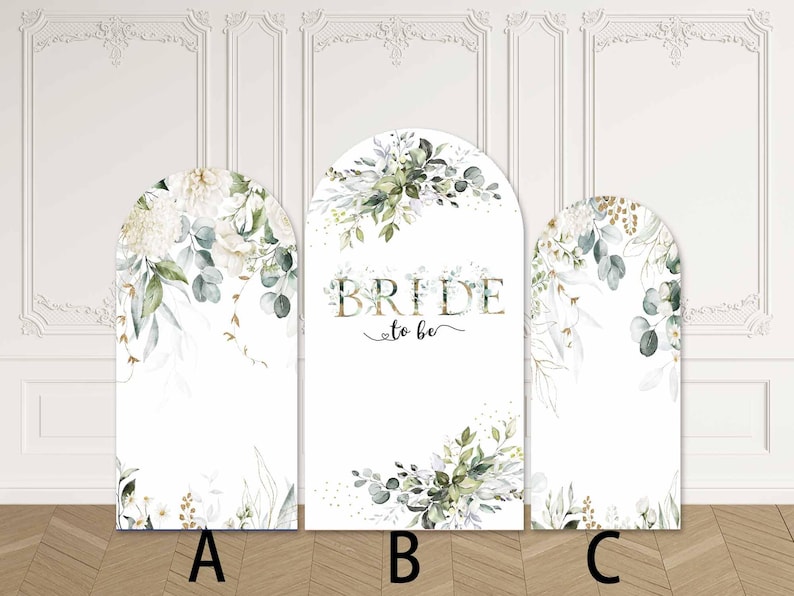May include: Three white arched panels with floral designs and greenery. The central panel reads "BRIDE to be" in gold lettering. The panels are decorated with white flowers, green leaves, and gold accents, ideal for a bridal shower or wedding.