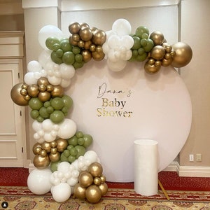 May include: A white round backdrop with gold lettering that reads "Dana's Baby Shower" decorated with a garland of white, green, and gold balloons.