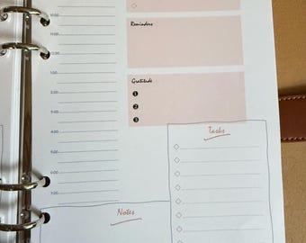 May include: Open planner with a daily schedule, top priorities, reminders, gratitude, tasks, and notes sections. The planner has a light brown cover and a metal ring binder. The text "Today is" and the days of the week are visible.