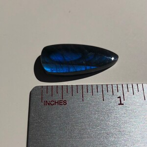 May include: A dark blue, iridescent, teardrop-shaped gemstone cabochon, measuring approximately 1 inch in length, is displayed on a ruler.