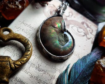 Puede incluir: Un collar con un colgante de plata con un fósil de amonita iridiscente. El colgante se coloca sobre un fondo de página de libro vintage con texto escrito a mano.