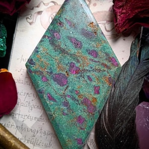 Könnte beinhalten: Ein rautenförmiger Edelstein mit einem gesprenkelten Muster aus Türkis, Magenta und Gold. Der Stein ist vor dem Hintergrund von getrockneten Rosen, Federn und einem antiken Schlüssel platziert, was auf ein mystisches oder böhmisches Thema hindeutet.