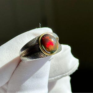 May include: A ring featuring a central oval-shaped dark opal with vibrant red and orange flashes, set in a gold-toned bezel. The band is crafted from textured silver-colored metal with a hammered finish and gold-toned linear accents on the sides.