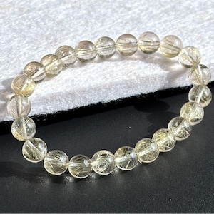 May include: A clear quartz bracelet with golden rutile inclusions. The round beads are strung together, creating a flexible, circular design. The bracelet is displayed on a black surface with a white, textured background.