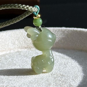 May include: A light green jade pendant in the shape of a deer. The deer pendant is suspended from a green cord with a small green bead and gold-colored accent. The pendant is smooth and polished, with a detailed deer design.