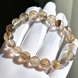 May include: A clear quartz bracelet with brown inclusions. The round beads are strung together, creating a circular design. The bracelet is displayed on a white surface, with light reflecting off the beads.