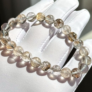 May include: A clear quartz bracelet with brown inclusions. The bracelet is made of round beads and is strung on a clear elastic cord.