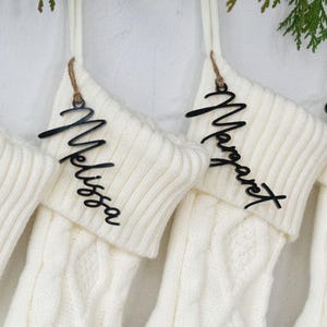 May include: Four cream-colored cable knit Christmas stockings hang from a white surface, each with a wooden name tag. The names on the tags are "Mommy", "Melissa", "Margaret", and "Elizabeth". Greenery is draped above the stockings.