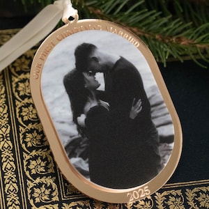 May include: Gold-toned Christmas ornament with a black and white photo of a couple kissing. The ornament has the text "OUR FIRST CHRISTMAS MARRIED" and the year "2025". A white ribbon is attached for hanging. The ornament is on a decorative book.