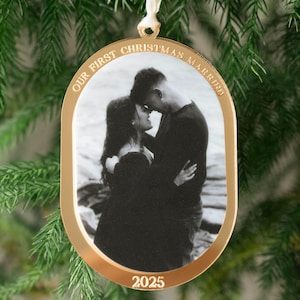 May include: Gold-colored Christmas ornament with a black and white photo of a couple embracing. The ornament has the text "OUR FIRST CHRISTMAS MARRIED" and "2025" in gold lettering. A white ribbon is attached for hanging.