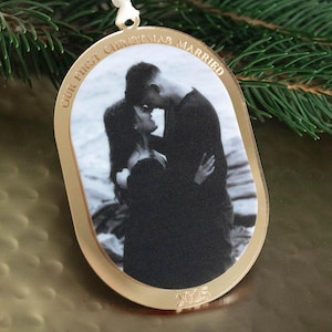 May include: Gold-colored oval Christmas ornament with a black and white photo of a couple embracing. The text "OUR FIRST CHRISTMAS MARRIED" is arched around the top edge, and the year "2025" is at the bottom. A white ribbon is attached for hanging.