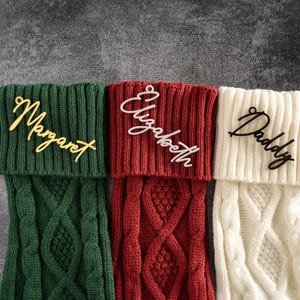 May include: Three personalized Christmas stockings in green, red, and cream. Each stocking features a cable knit design and a name: "Margaret" in gold, "Elizabeth" in white, and "Daddy" in black. The stockings are displayed against a gray background.