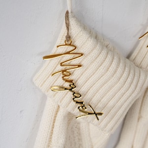 May include: A gold-colored acrylic name tag that says "Margaret" in a cursive font, hanging from a cream-colored knitted stocking. The tag is suspended by a thin, natural-colored twine. The stocking has a ribbed knit texture.