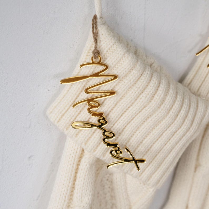 May include: A gold-colored acrylic name tag that says "Margaret" in a cursive font, hanging from a cream-colored knitted stocking. The tag is suspended by a thin, natural-colored twine. The stocking has a ribbed knit texture.