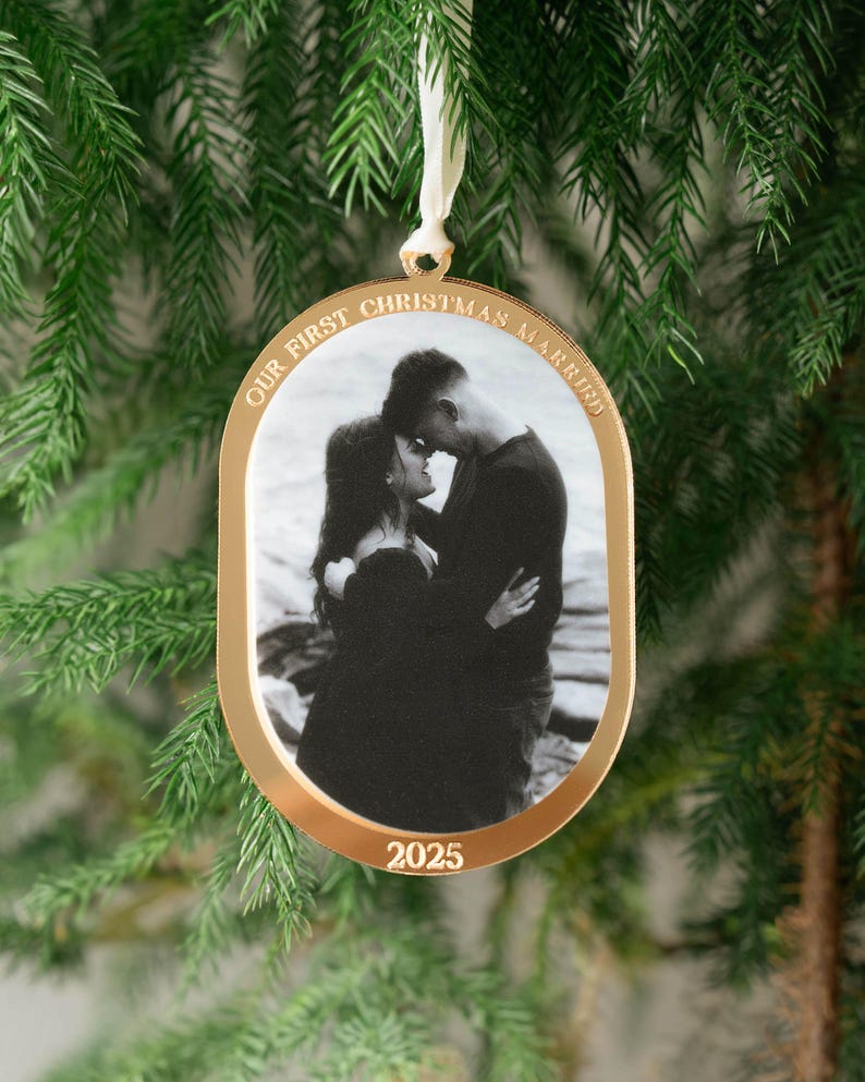May include: Gold-colored Christmas ornament with a black and white photo of a couple embracing. The ornament has the text "OUR FIRST CHRISTMAS MARRIED" and "2025" in gold lettering. A white ribbon is attached for hanging.