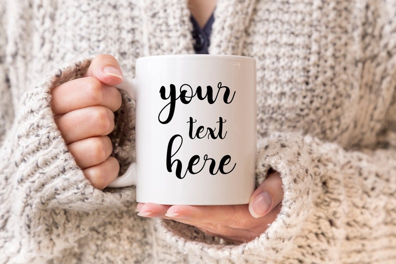 May include: White ceramic mug with black cursive text that reads "your text here". The mug is being held by a person wearing a beige sweater.