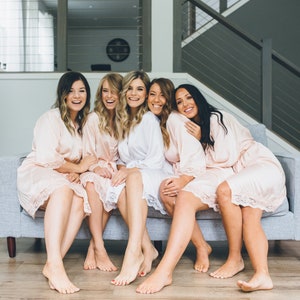May include: Five women wearing pink and white robes are sitting on a gray couch. They are smiling and looking at the camera. The couch is in a room with a wooden floor and a staircase in the background.