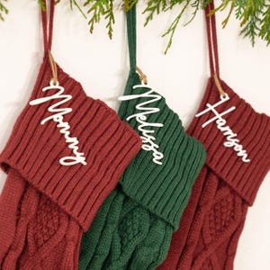 May include: Three Christmas stockings in red and green hang from a branch. Each stocking has a white name tag: "Mommy", "Melissa", and "Harrison". The stockings are made of a knitted material.