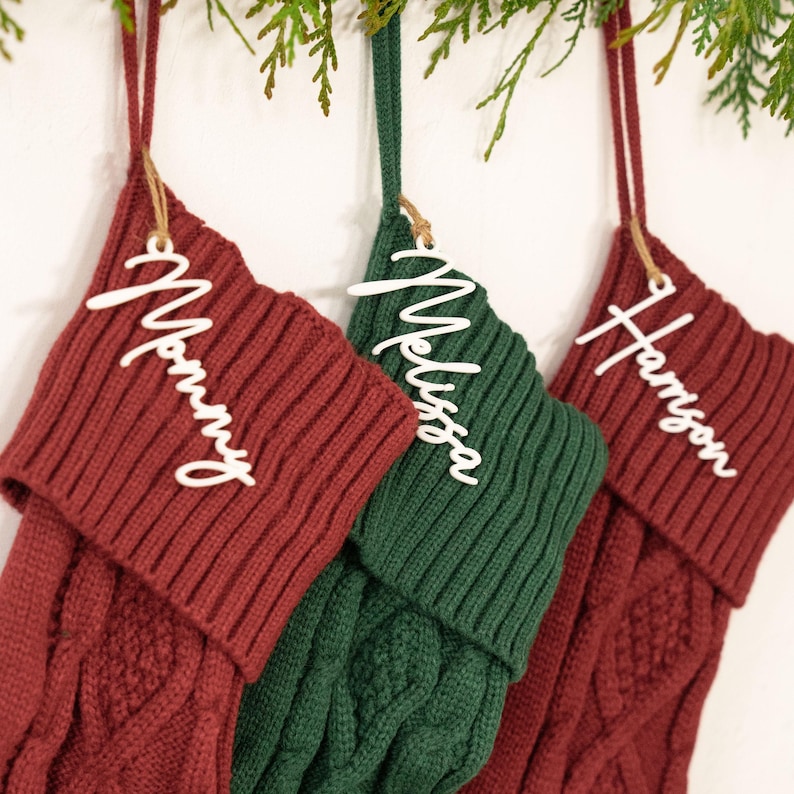 May include: Three Christmas stockings in red and green hang from a branch. Each stocking has a white name tag: "Mommy", "Melissa", and "Harrison". The stockings are made of a knitted material.