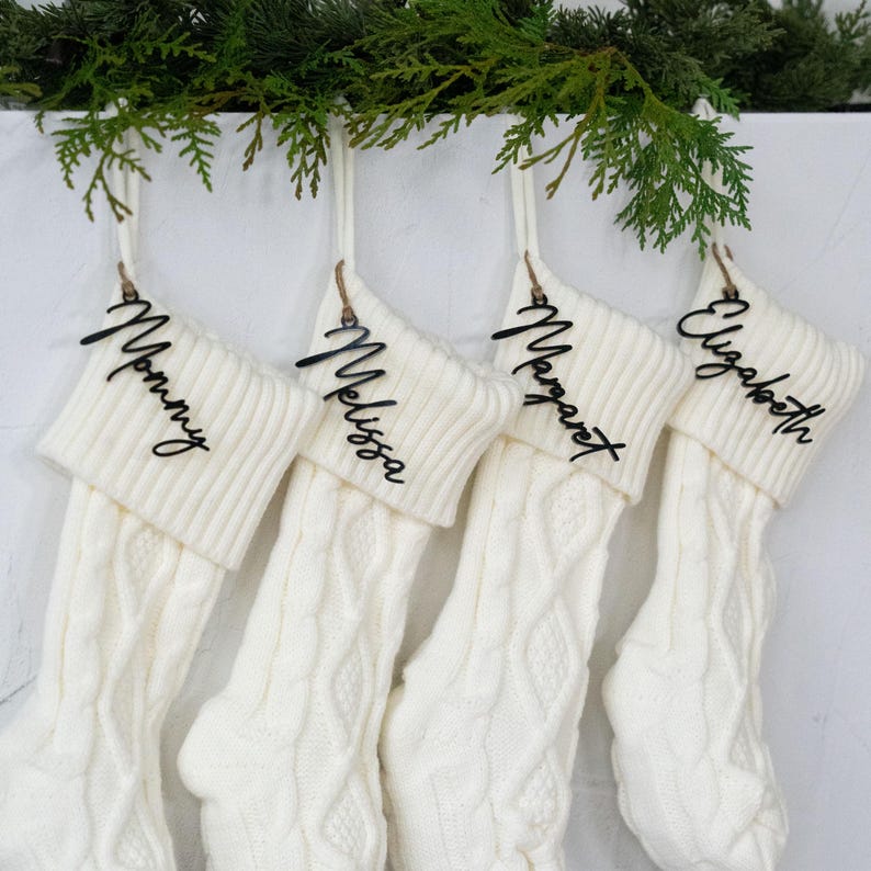 May include: Four cream-colored cable knit Christmas stockings hang from a white surface, each with a wooden name tag. The names on the tags are "Mommy", "Melissa", "Margaret", and "Elizabeth". Greenery is draped above the stockings.