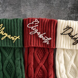 May include: Three Christmas stockings in green, red, and cream, each with a nameplate. The green stocking has "Margaret" in gold script, the red has "Elizabeth" in white, and the cream has "Daddy" in black script. The stockings have a cable knit design.