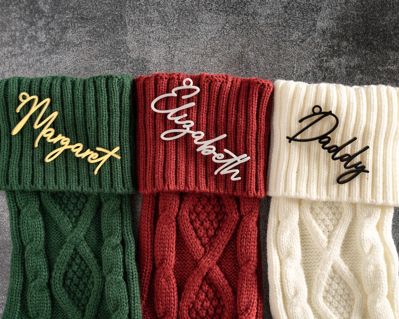 May include: Three Christmas stockings in green, red, and cream, each with a nameplate. The green stocking has "Margaret" in gold script, the red has "Elizabeth" in white, and the cream has "Daddy" in black script. The stockings have a cable knit design.