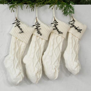 May include: Four off-white cable knit Christmas stockings hang from a green garland. Each stocking has a black name tag that reads "Mommy", "Melissa", "Margaret", and "Elizabeth". The stockings are suspended against a white background.