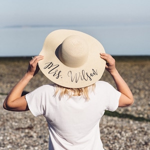 Puede incluir: Una mujer con una camiseta blanca y pantalones vaqueros azules lleva un sombrero de paja para el sol con el texto "Mrs. Wilson" escrito en &eacute;l.